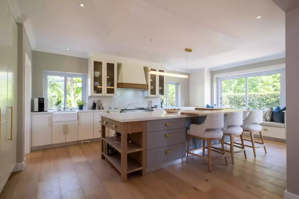Elegant kitchen featuring a large center island with integrated grey drawers, a wood block end piece, light grey upholstered bar stools, white perimeter cabinets with gold handles, and a window bench seat.