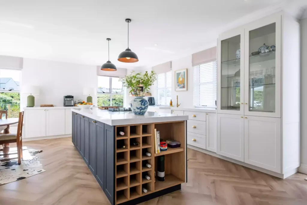 Spacious kitchen featuring a large black center island with a white marble countertop, a built-in wine rack and wooden open shelving, black pendant lights, a large glass-front display cabinet, and herringbone hardwood flooring.