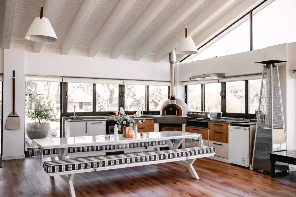 A modern indoor braai and entertainment area featuring a white picnic-style dining table with striped bench cushions, an integrated wood-fired pizza oven, built-in cabinetry, and a high vaulted ceiling with white beams.