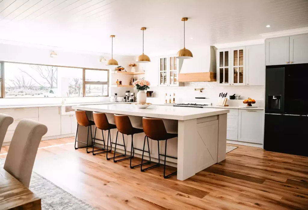 A bright farmhouse kitchen featuring a large white island with four brown leather counter stools, three gold dome pendant lights hanging from a white shiplap ceiling, white cabinetry, a black refrigerator, and warm wood flooring.