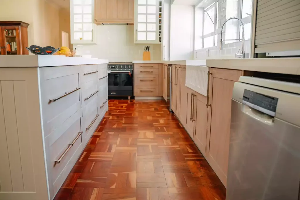 A bright traditional kitchen featuring light-toned wooden cabinetry with slim metal handles, white solid countertops, and classic reddish-brown parquet wood flooring. The layout includes a central island with deep drawers on the left, a long counter with a white ceramic butler sink and a goose-neck mixer tap under a window on the right, and a black freestanding gas stove beneath a wooden extractor hood in the background.
