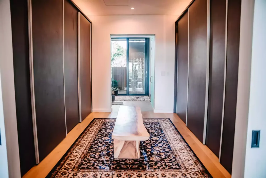 A walk-in closet or dressing room featuring floor-to-ceiling dark wooden sliding wardrobe doors lining both sides of the passage. A traditional patterned area rug sits on the light wood flooring, topped with a rustic, solid wood bench, leading toward a glass door opening to an outdoor patio.