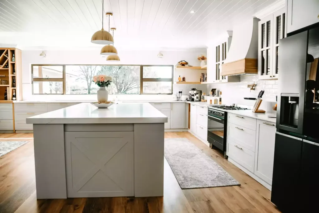 A large farmhouse-style kitchen featuring a white central island with an X-brace detail, white cabinetry with gold handles, gold dome pendant lights, a black stove, and a wide window looking out to trees.