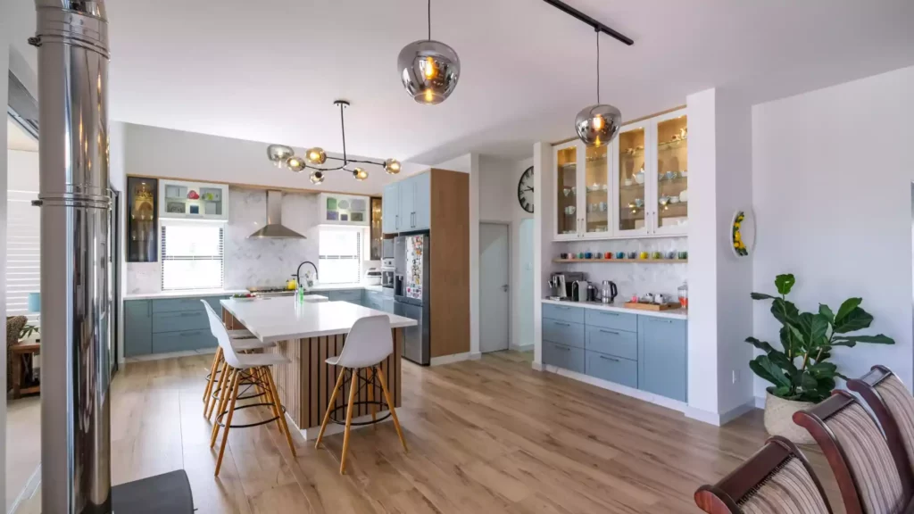 Bright and spacious modern open-plan kitchen featuring a large central island with seating, light blue and wood cabinetry, glass display cabinets, and spherical pendant lighting over wood floors.