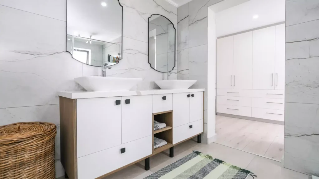 Modern bathroom vanity with white cabinets, double vessel sinks, and unique shaped mirrors against a marble tile wall, leading into a white walk-in closet.