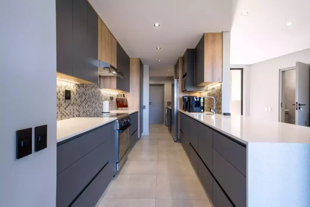 Modern galley kitchen with dark grey handleless cabinets, light wood accents, a white countertop island, a built-in stainless steel stove, and a grey hexagon tile backsplash.