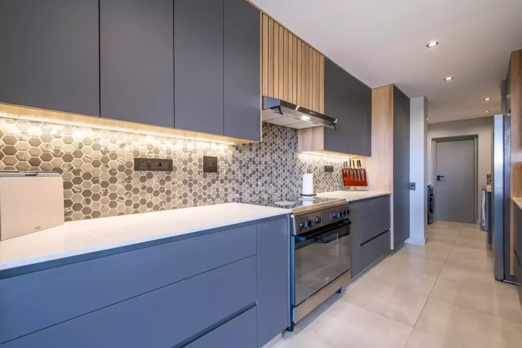 Modern kitchen with matte grey handleless cabinets, a freestanding stainless steel gas stove, a white countertop, under-cabinet lighting, and a grey hexagon mosaic tile backsplash.