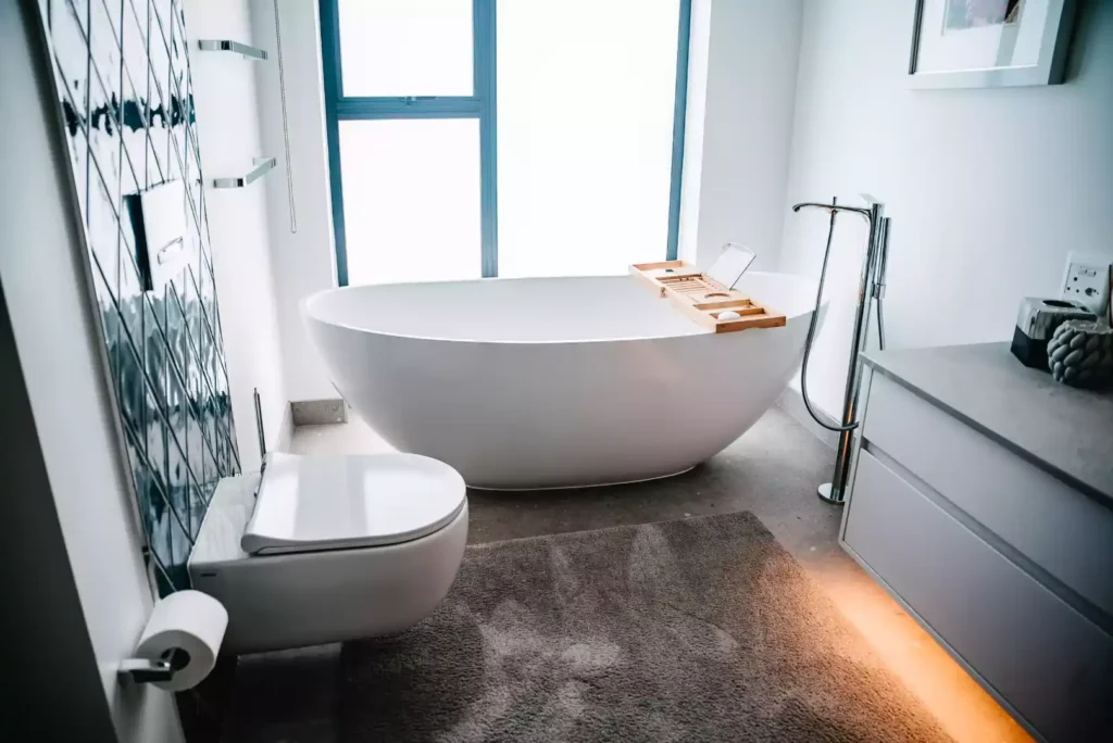 A modern bathroom featuring a freestanding white oval bathtub with a wooden caddy tray, a wall-mounted floating toilet, a grey vanity with under-cabinet lighting, a brown plush bath mat, and a large window in the background.