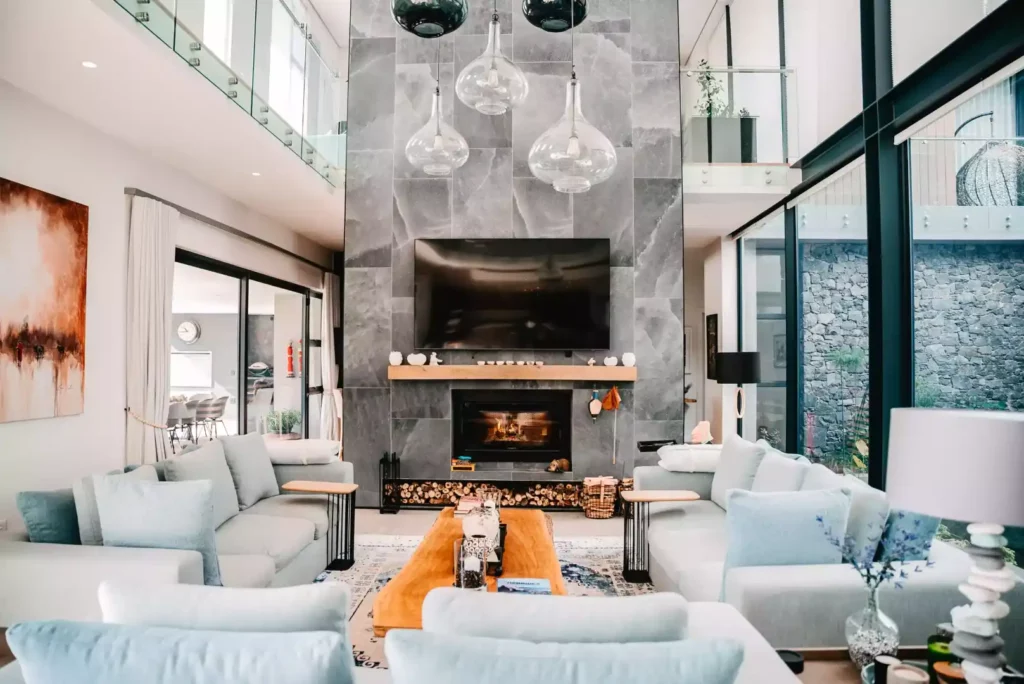 A luxurious double-volume living room featuring a massive grey stone fireplace wall with a built-in television, wood storage at the base, and hanging glass pendant lights. The space is furnished with matching light grey sofas, a solid wood coffee table, and large glass windows overlooking a patio.