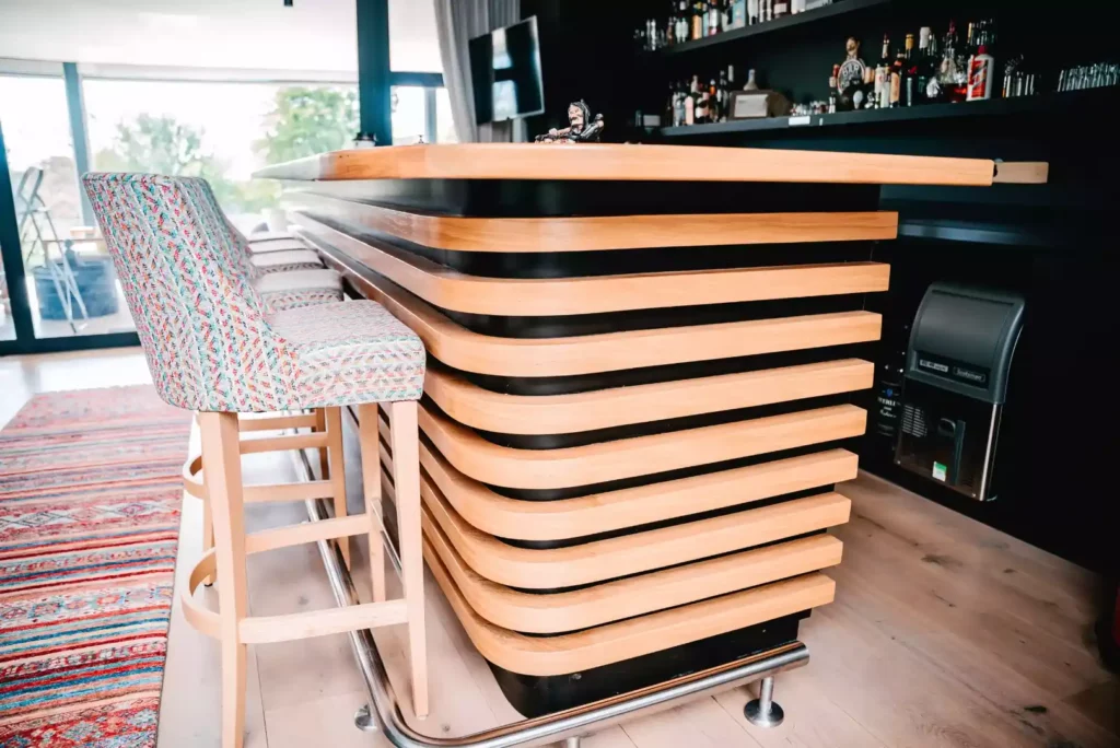 A modern home bar featuring a curved counter front detailed with horizontal light wood slats over black backing, paired with wooden bar stools upholstered in a multi-colored patterned fabric, set against a black wall with liquor shelves.