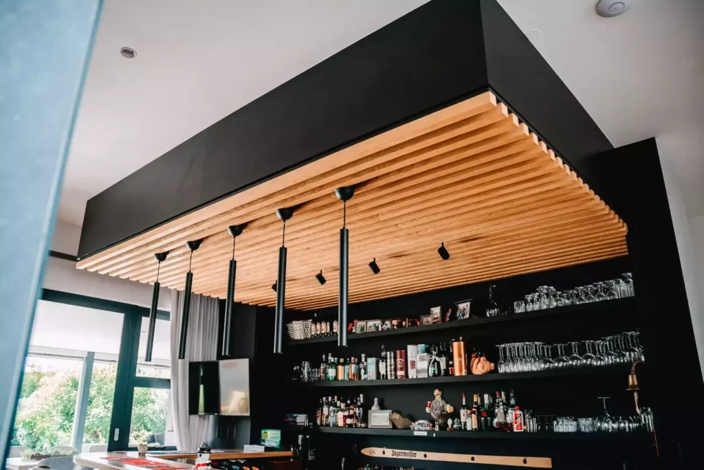 A modern home bar featuring an architectural dropped ceiling element with vertical light wood slats and a matte black border, long black cylindrical pendant lights, and backlit black shelves stocked with liquor bottles and glassware.