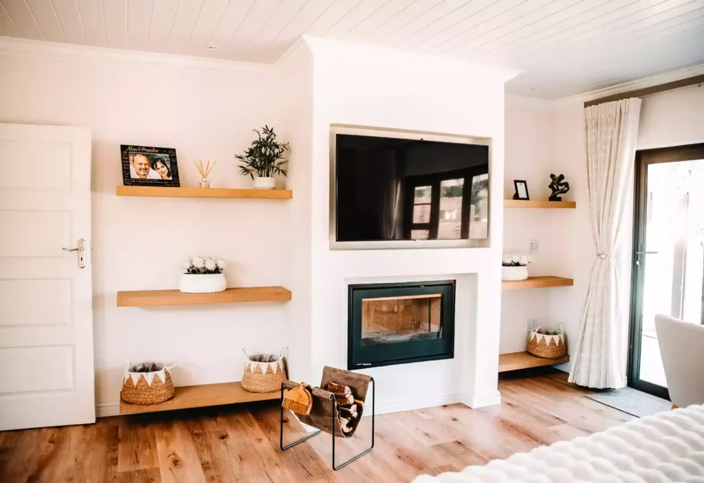 A modern living room space featuring a central white partition wall with a recessed TV mounted above a built-in black fireplace, flanked by light wood floating shelves displaying decor and woven baskets.