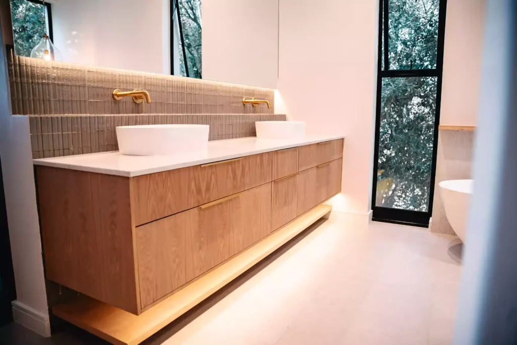 A long walk-in closet featuring light wood cabinetry with white countertops, open hanging rods filled with clothes, woven storage baskets on top shelves, a central upholstered bench, and a large window at the far end.