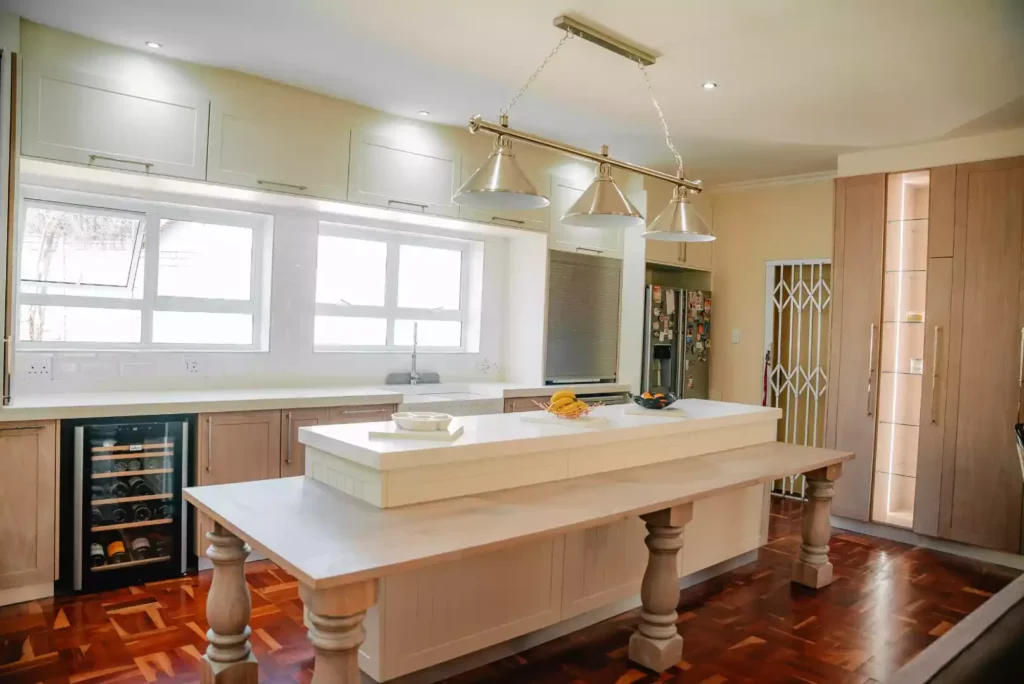 A wide view of a bright traditional kitchen featuring a central island with a raised white countertop and a lower long wooden breakfast table extension supported by thick turned legs. Above the island hangs a modern linear three-lamp silver pendant light fixture. The space includes classic reddish-brown parquet wood flooring, light wood cabinetry, an under-counter wine fridge, white tiled splashbacks under wide windows, and tall built-in wood-paneled storage units in the background.