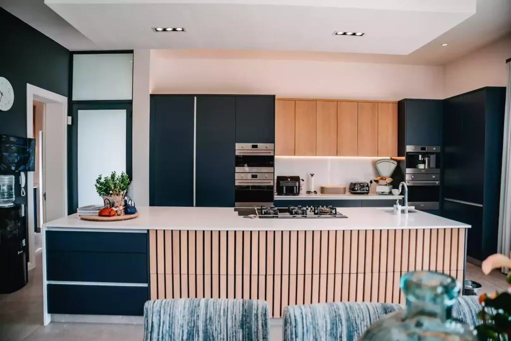 A straight-on view of a modern kitchen featuring a large central island with a white countertop, an integrated gas stovetop, and a front detailed with vertical light wood slats and dark charcoal side drawers. The back wall includes dark charcoal cabinetry, light wood upper cabinets with under-cabinet lighting, and integrated stainless steel ovens.