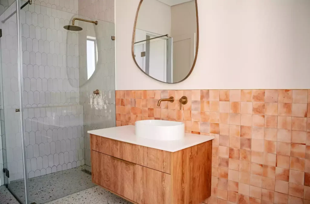 A modern bathroom featuring a floating wooden vanity with vertical slat details, a white vessel sink with a wall-mounted gold tap, an organic-shaped gold-framed mirror, and a wall lined with square terracotta tiles. On the left, a glass-enclosed walk-in shower showcases white scalloped tiles, a gold rain shower head, and terrazzo flooring.