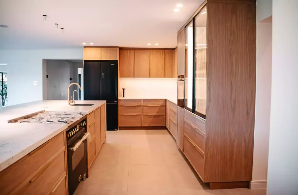 A modern kitchen featuring warm medium-wood cabinetry, a large central island with a white marble countertop, a built-in black gas range oven, and a gold goose-neck tap. The back wall includes a black French-door refrigerator and a tall pantry unit with internal lighting and reeded glass doors.