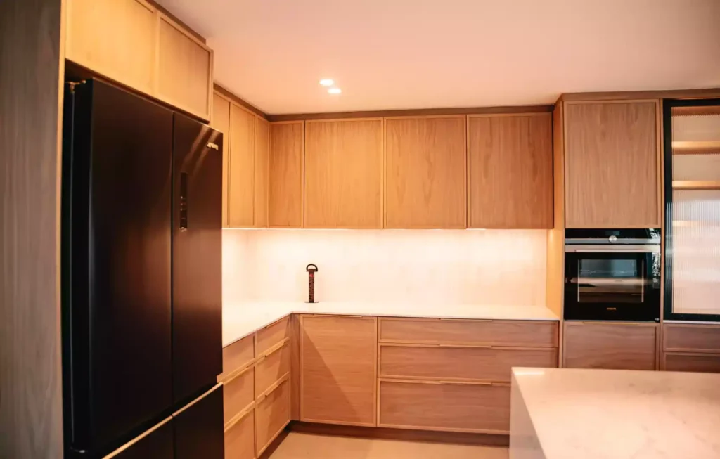 A modern kitchen corner featuring warm medium-wood cabinetry with under-cabinet LED lighting, a black French-door refrigerator on the left, white countertops, and an integrated wall oven on the right.