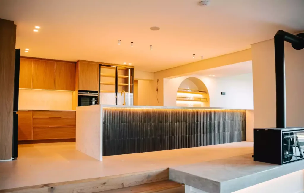 A modern kitchen featuring a massive marble waterfall island with a dark, vertically textured tile front illuminated by an under-counter LED strip light. The background displays warm medium-wood cabinetry, glass-door pantry storage, and an arched wall niche, while a black freestanding fireplace sits on a concrete plinth in the foreground.