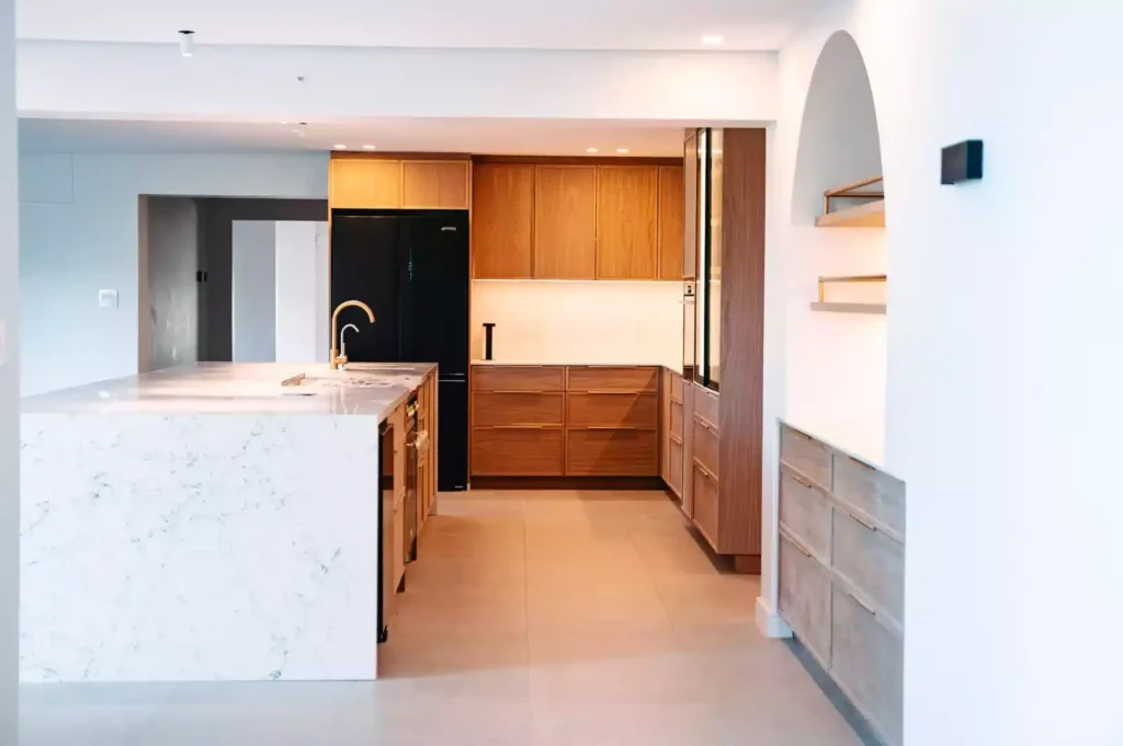 A modern kitchen featuring a large marble waterfall island with a gold prep faucet, warm medium-wood cabinetry, a black French-door refrigerator, and an arched wall niche with integrated open shelving and a matching wood buffet counter.