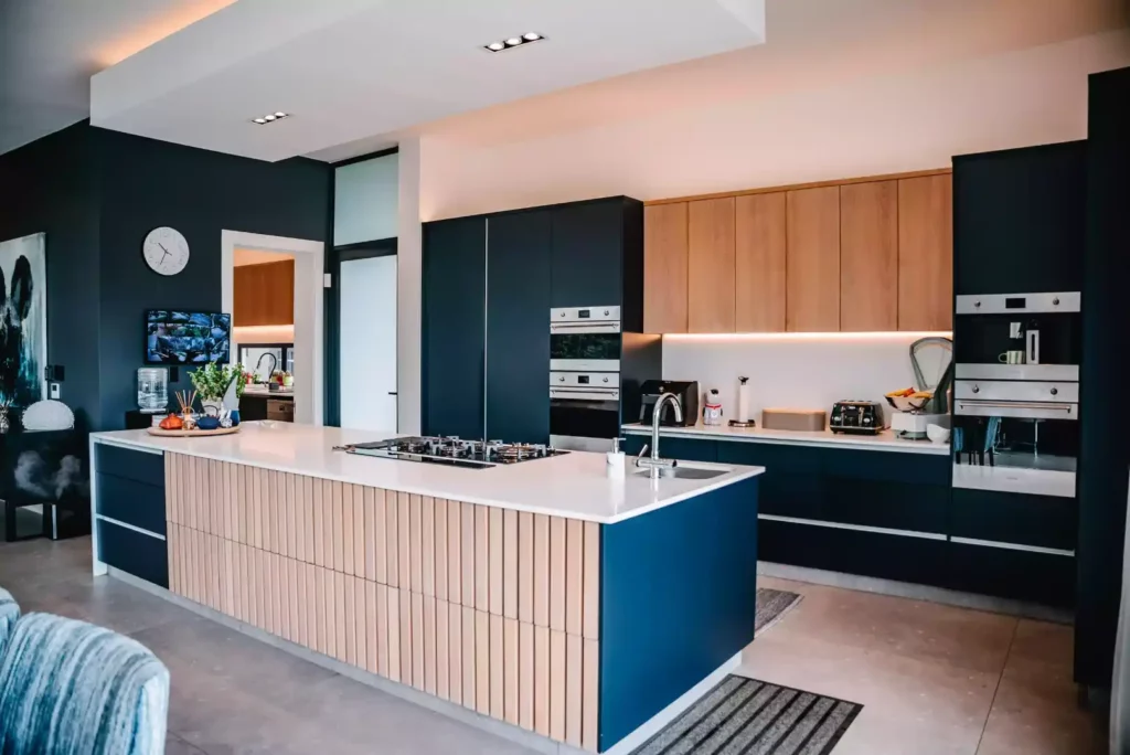 A modern kitchen featuring a large central island with a white countertop, an integrated gas stovetop, a prep sink, and a front detailed with vertical light wood slats and deep blue end panels. The back wall is lined with dark charcoal cabinetry, light wood upper cabinets with under-cabinet LED lighting, and integrated stainless steel wall ovens.