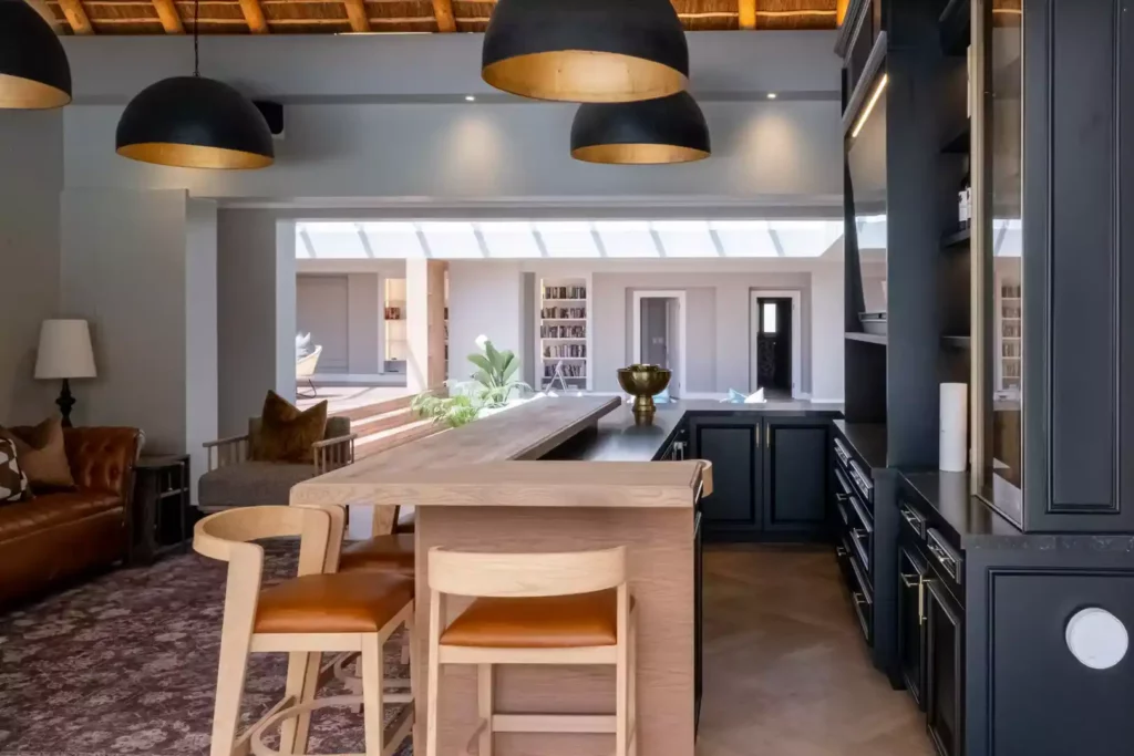 Luxurious home bar and entertainment area featuring a thick wooden L-shaped bar counter with leather-seat bar stools, black cabinetry, large black dome pendant lights with gold interiors, and a view into an adjacent open living space under a thatched roof.