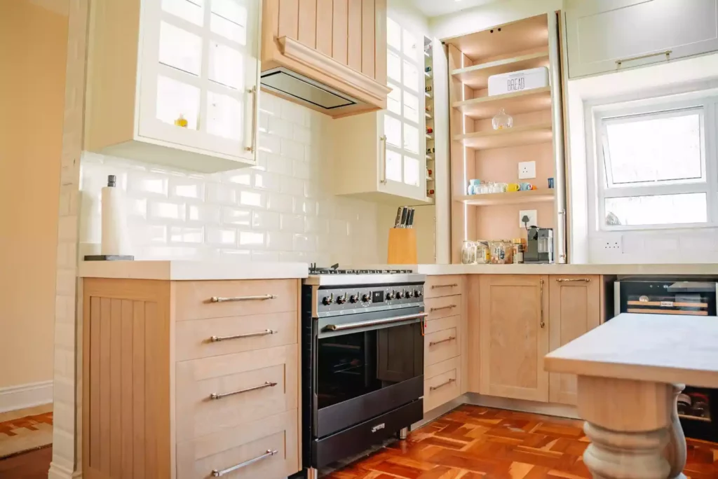 A bright, traditional kitchen featuring light-toned wooden cabinetry with slim silver handles, white solid countertops, and classic reddish-brown parquet wood flooring. In the center sits a large black freestanding gas range cooker beneath a wooden extractor hood, flanked by white tiled splashbacks and upper cabinets with grid-glass doors. To the right, a tall built-in pantry unit with open wooden shelving holds kitchen items like a bread bin and coffee machine, next to a small under-counter wine fridge.