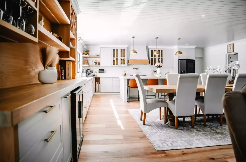 A bright, open-concept kitchen and dining area featuring a wooden bar station in the foreground, a large wooden dining table with upholstered grey chairs on a patterned rug, and a farmhouse-style kitchen with white cabinetry and gold pendant lights in the background.