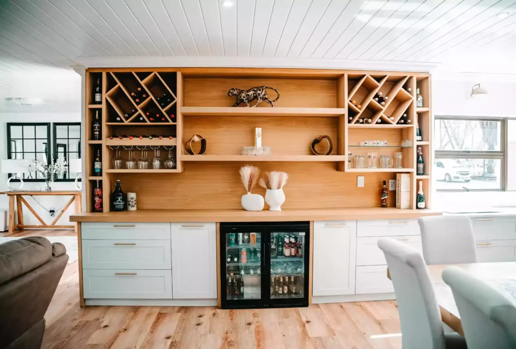A modern home bar and beverage station featuring a large wooden upper unit with built-in diamond wine racks, open display shelving, and under-cabinet glass holders, set above white lower cabinetry with a built-in double-door beverage refrigerator.