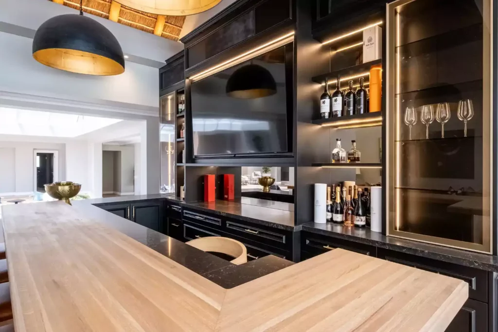 Luxurious home bar featuring black cabinetry, integrated LED strip lighting, a built-in TV screen, mirrored back panels, a glass display case with wine glasses, and a thick wooden L-shaped bar counter.
