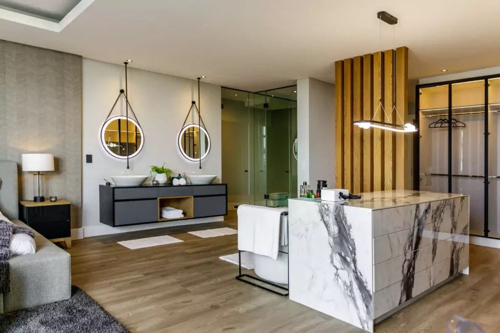 Luxury open-plan master bedroom and en-suite bathroom featuring a double black floating vanity, backlit hanging circular mirrors, a marble island dresser, and a glass-enclosed shower.