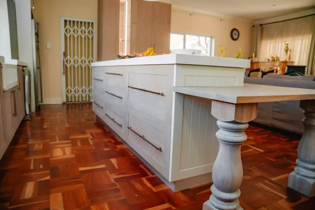 A bright, traditional kitchen featuring a central island with a white marble countertop, light-toned wooden drawers, and a built-in lower wooden breakfast bar extension supported by thick, ornate turned wooden legs. The space includes a classic reddish-brown parquet wood floor, a security gate in the background, and a glimpse of an open-plan living area beyond the island.