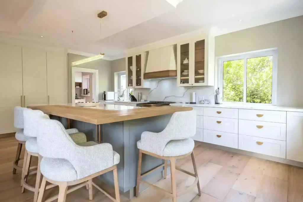 Elegant kitchen featuring a large grey center island with a wood overlay countertop, light grey upholstered bar stools, white perimeter cabinets with half-moon brass handles, and glass-front upper cabinets.