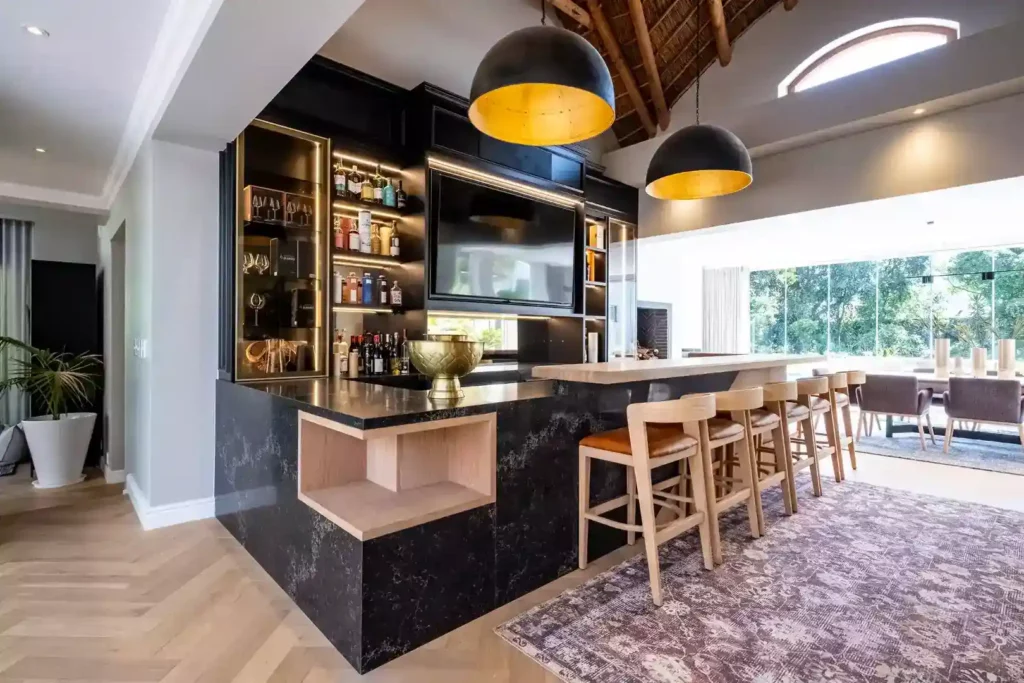 Luxurious open-plan home bar featuring a black marble counter with an open wood display nook, lined with wooden bar stools with brown leather seats, black back cabinetry with integrated lighting and a built-in TV screen, and large black dome pendant lights with gold interiors under a thatched roof.