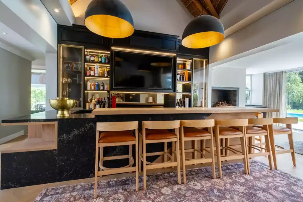Luxurious home bar featuring a black marble counter topped with a thick wooden overlay, lined with wooden bar stools with brown leather seats, set against black cabinetry with integrated LED strip lighting, a built-in TV, and large black dome pendant lights with gold interiors.