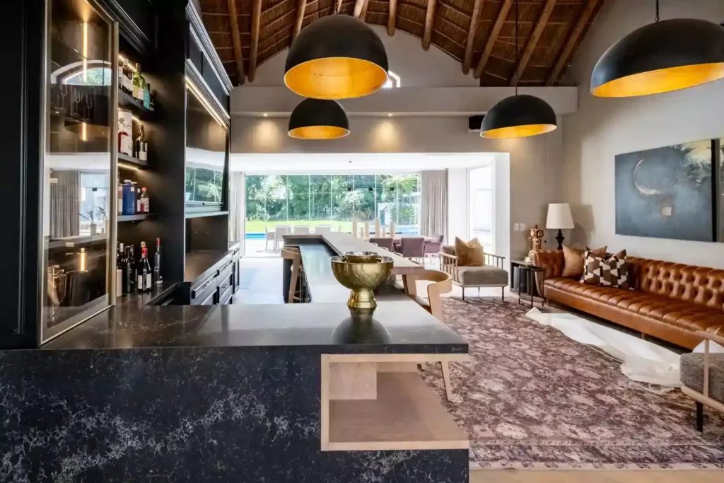 Luxurious open-plan entertainment area with a high thatched roof, featuring a black marble bar counter, large black dome pendant lights with gold interiors, a brown leather Chesterfield sofa, and an expansive view of the outdoor patio and garden.