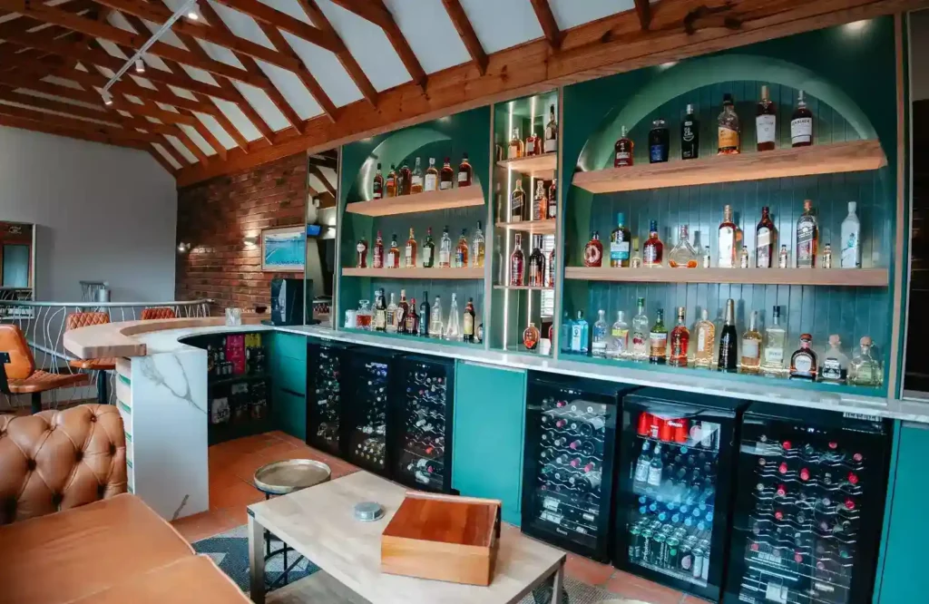 A wide view of a spacious rustic home bar featuring a long back counter with teal cabinetry and multiple illuminated arched shelving bays filled with liquor bottles. Five glass-front beverage and wine fridges are integrated beneath a white marble countertop. The bar setup includes a curved wooden front counter with marble detailing, a low wooden coffee table in the foreground, tufted brown leather seating, and an exposed brick accent wall under a high vaulted wooden ceiling.