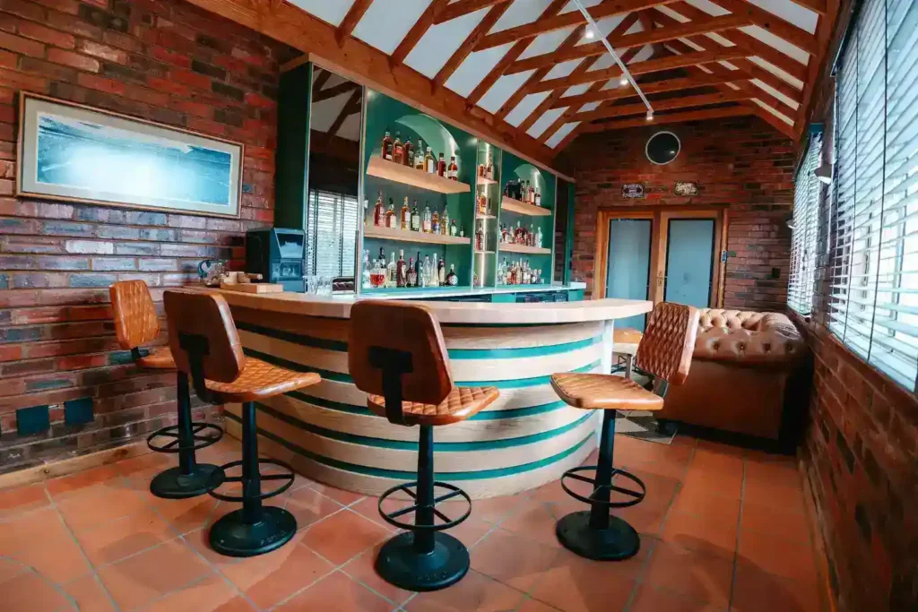 A rustic home bar room featuring a curved wooden front counter with tiered slat details and green painted accents. Four brown quilted leather bar stools sit on a terracotta tile floor facing the bar. The back unit consists of teal cabinets and illuminated arched shelves displaying various liquor bottles. The entire space is framed by exposed brick walls, window blinds on the right, and a high vaulted wooden ceiling with exposed rafters.