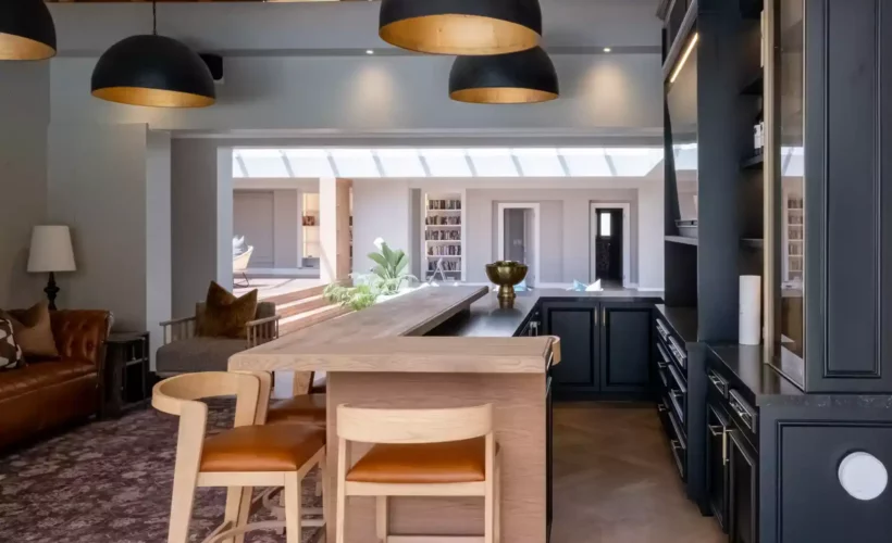 Luxurious home bar and entertainment area featuring a thick wooden L-shaped bar counter with leather-seat bar stools, black cabinetry, large black dome pendant lights with gold interiors, and a view into an adjacent open living space under a thatched roof.