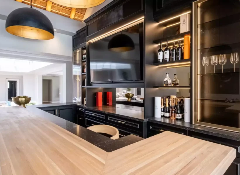 Luxurious home bar featuring black cabinetry, integrated LED strip lighting, a built-in TV screen, mirrored back panels, a glass display case with wine glasses, and a thick wooden L-shaped bar counter.